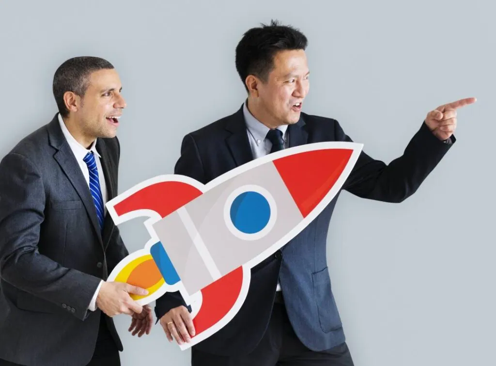 Two men in business suits, one holding a large cutout of a red rocket, the other pointing forward, both looking in the same direction.