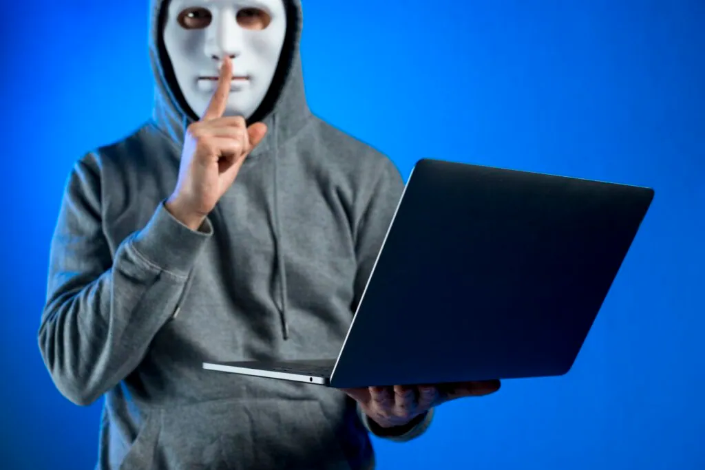 Person in a mask holding a laptop with a shushing gesture