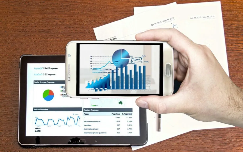 Hand holding smartphone showing financial graphs over tablet